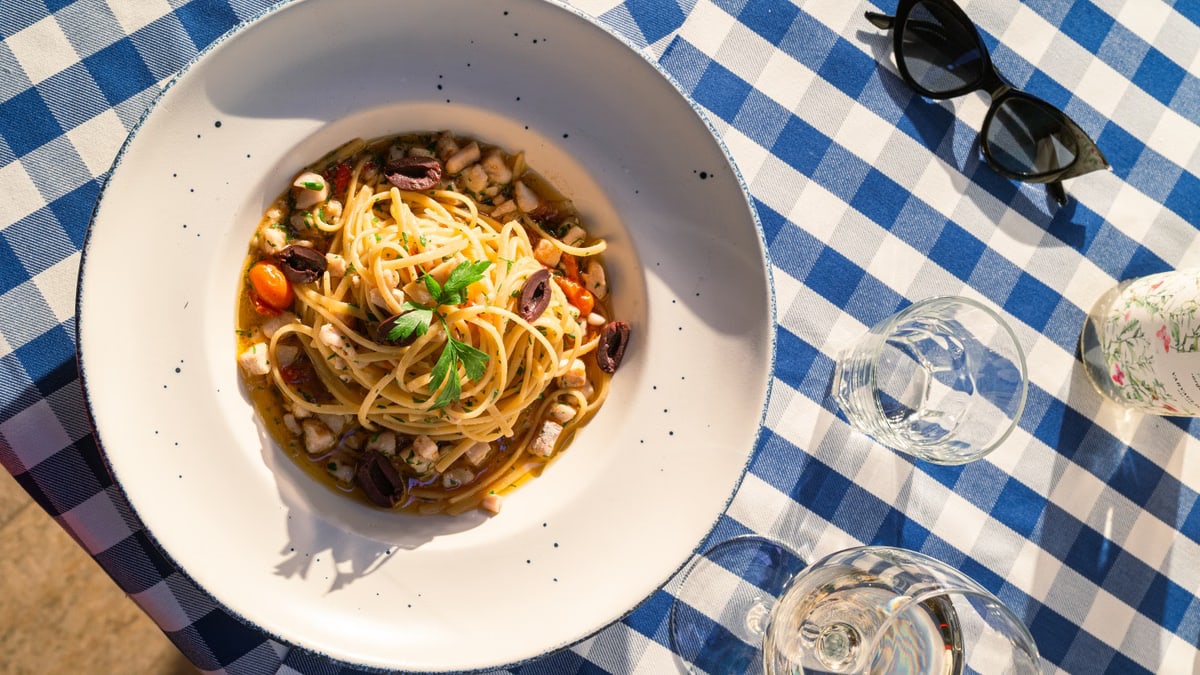 Top view of Italian pasta with parsley and olives on a blue checkered tablecloth with glasses in sunlight.